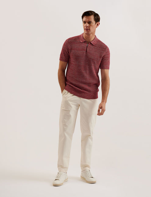 A male model wearing a red short sleeve polo shirt with a classic collar and a zip neck, paired with white trousers and white shoes.
