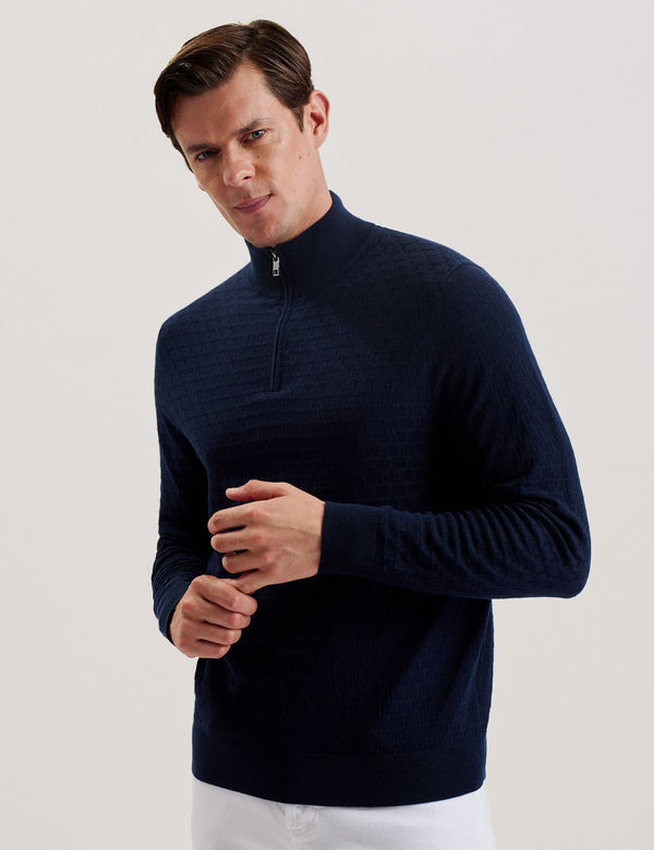 A man wearing a navy long sleeve wool blend jumper with a funnel neck and half zip placket, paired with white trousers.