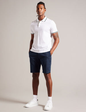 A man standing in a studio setting, wearing navy knee-length chino shorts with belt loops and pockets, paired with a white polo shirt and white sneakers.