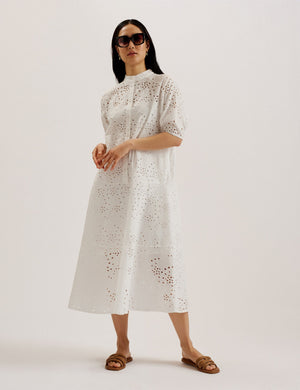 A woman wearing a white midi dress with broderie anglaise design, featuring an oversized fit, puff half sleeves, and a half button placket with a grandad collar.